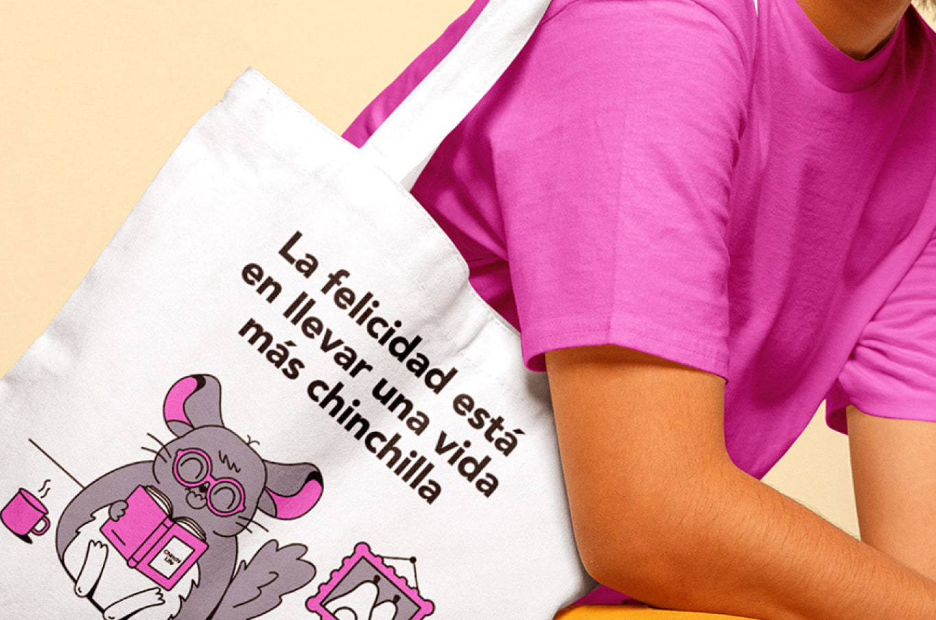 A close crop of a white canvas Cornershop tote carried by someone in a pink t-shirt against a pale yellow backdrop, printed with a chinchilla in glasses reading in an armchair under the line “La felicidad está en llevar una vida más chinchilla”.