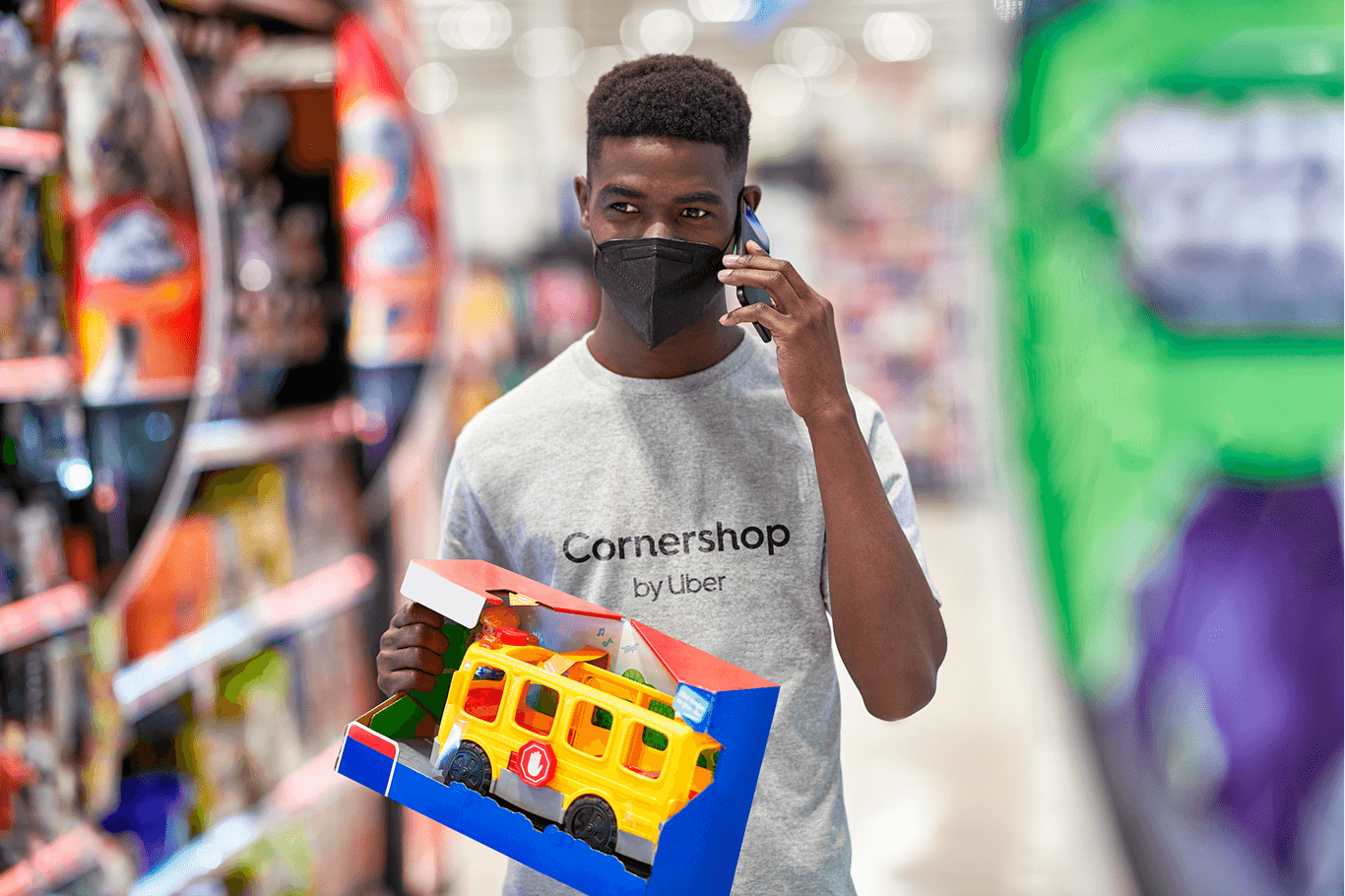A Cornershop shopper in a supermarket aisle, on the phone, holding a boxed toy bus. His t-shirt reads Cornershop, by Uber.
